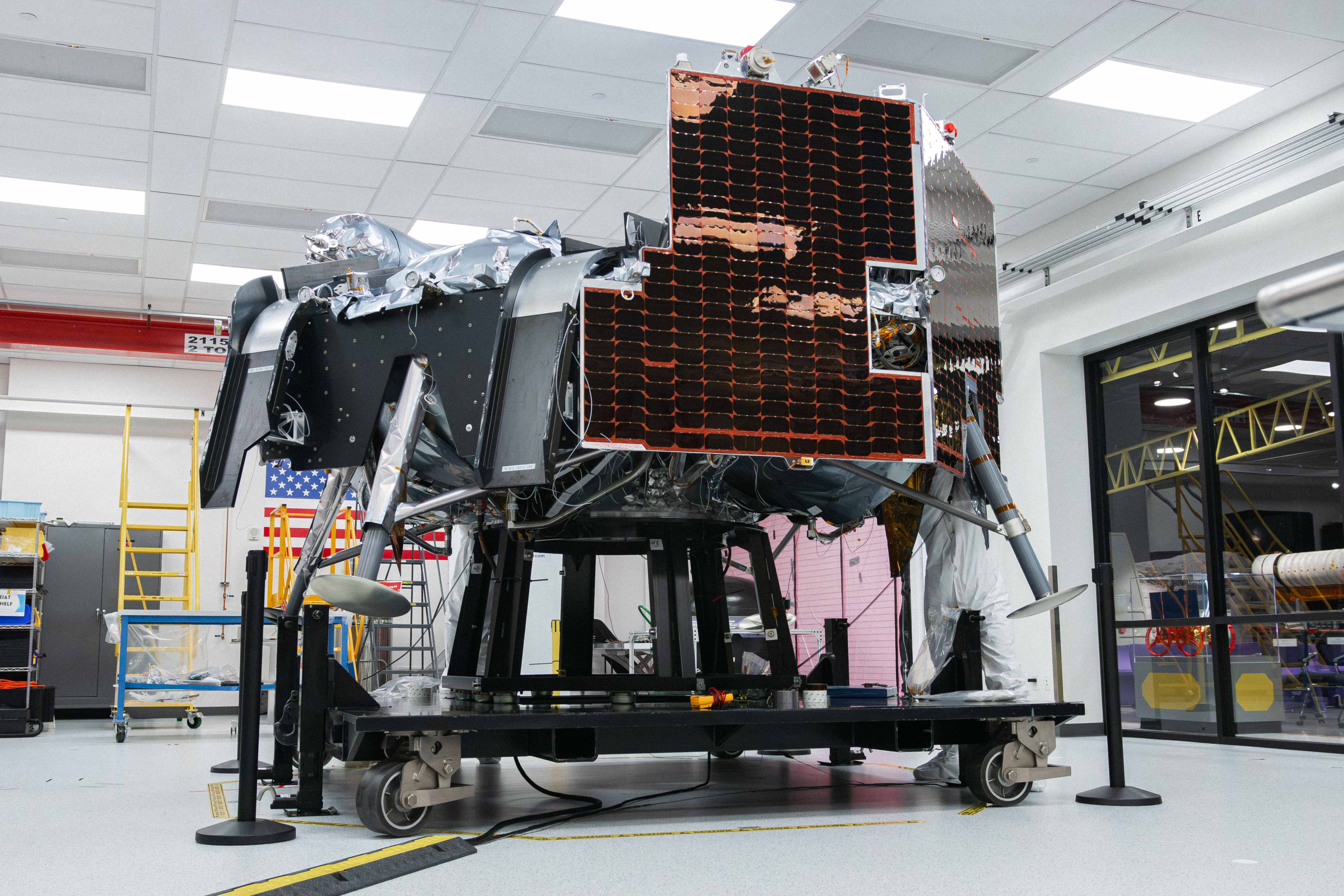 Astrobotic’s Griffin-1 lunar lander sits inside a clean room at the company’s facility. The lander is covered in dark solar panels and mounted on a wheeled test stand. Engineers in white clean-room attire work nearby, with the Astrobotic logo and a large American flag visible in the background.