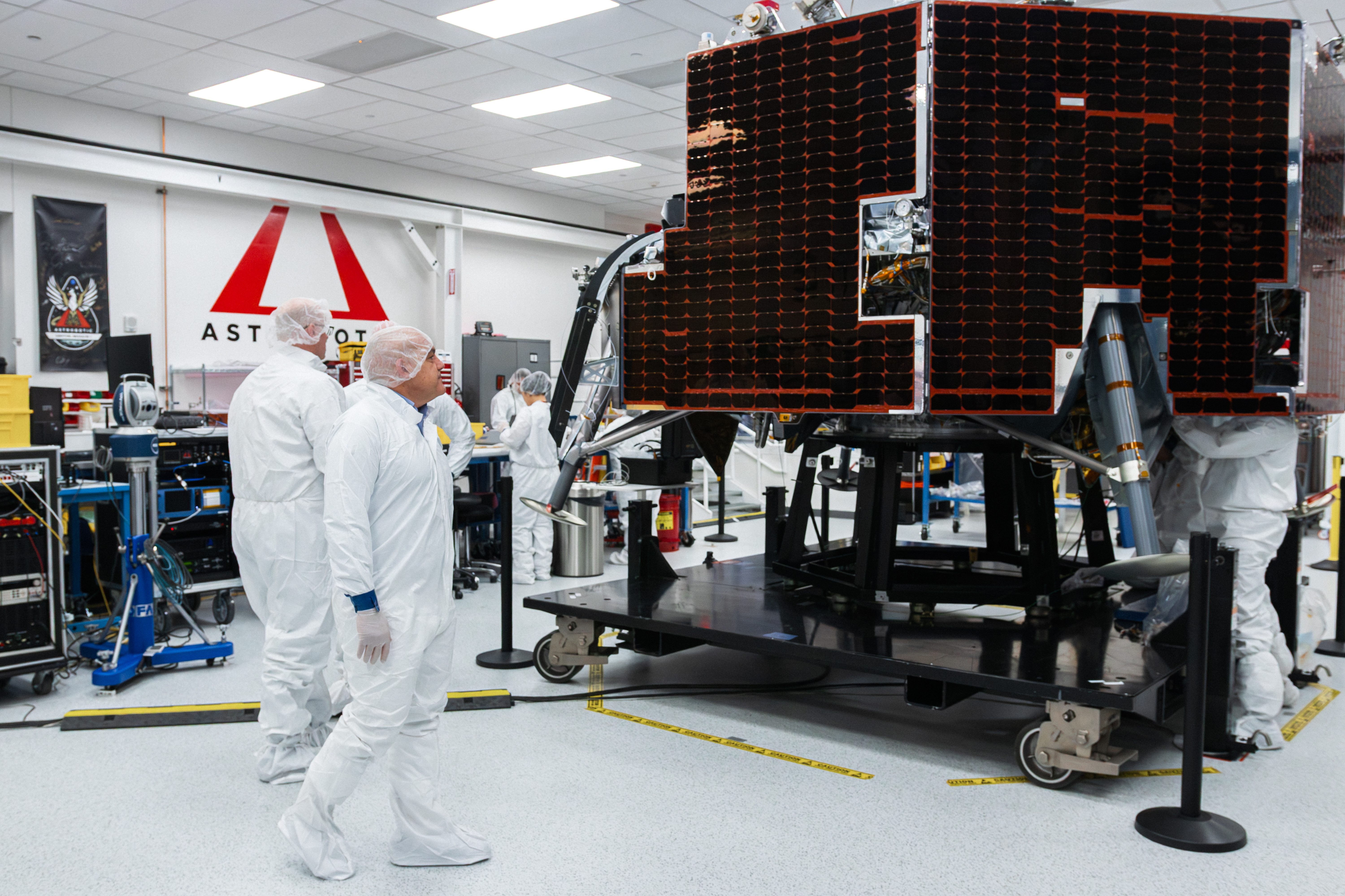 NASA Moon Base program leaders walk around Astrobotic’s Griffin-1 lunar lander inside a clean room in Pittsburgh, Pennsylvania. Large dark solar panels covers the side of the spacecraft as engineers work nearby in the background.