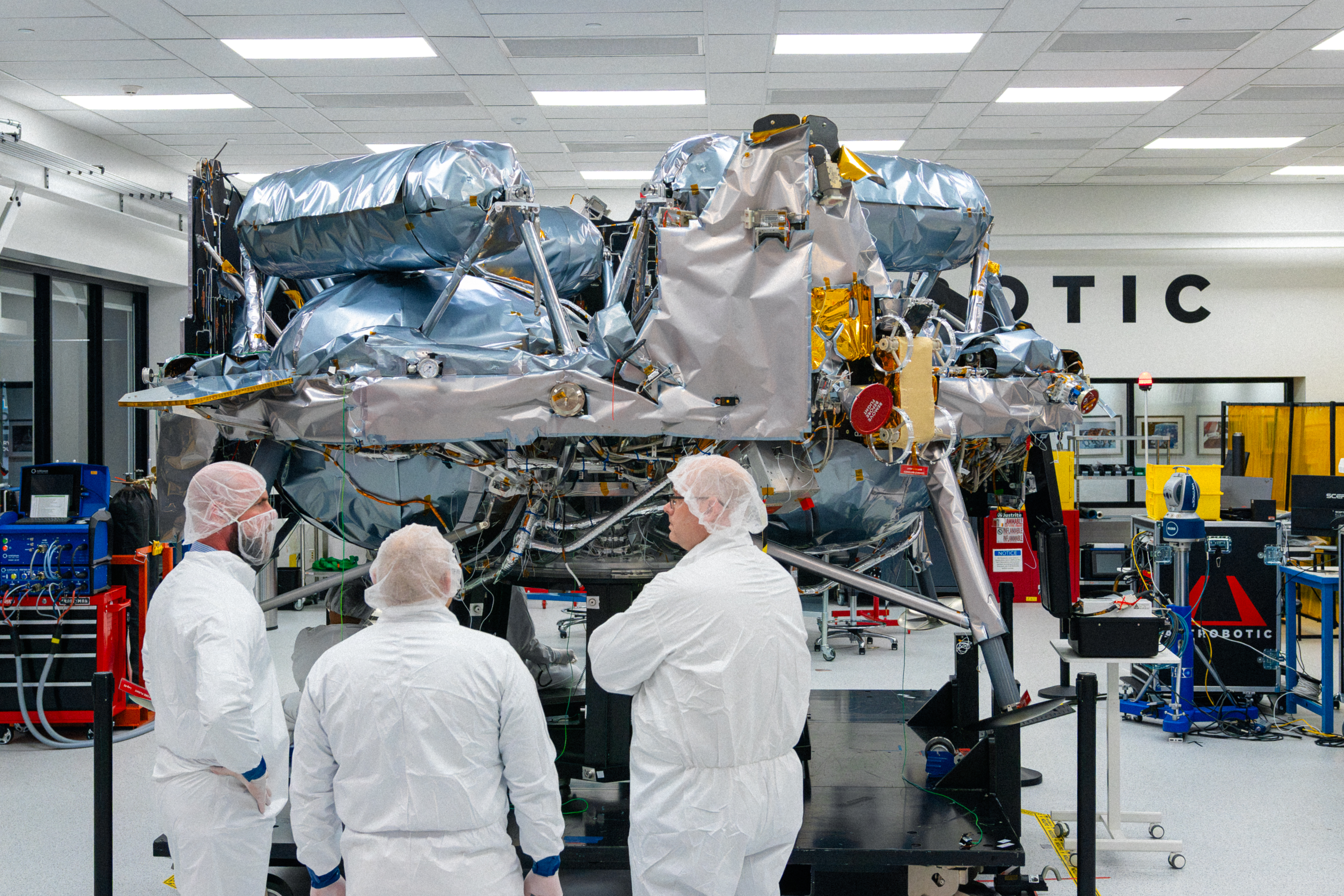 Three people stand in front of Astrobotic’s Griffin-1 lunar lander inside a clean room in Pittsburgh, Pennsylvania. The large lunar lander dominates the foreground as the group views the spacecraft.