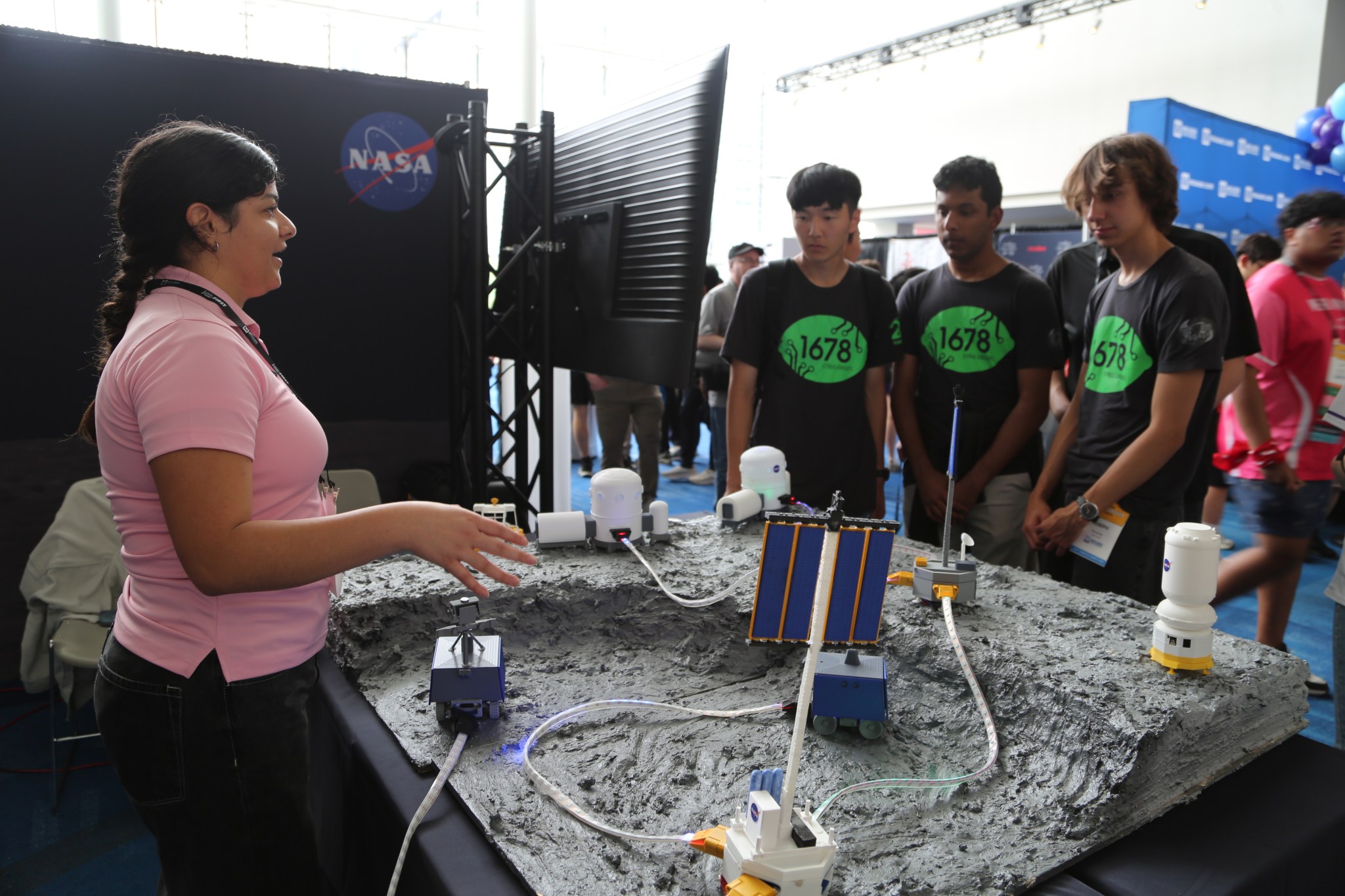A woman wearing a pink polo shirt explains a model of NASA's Moon Base to a three male high school students wearing matching t-shirts.