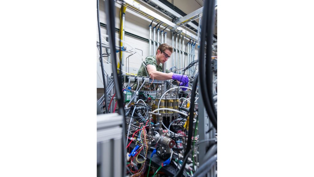 A researcher wearing purple gloves, safety glasses, and a green plaid shirt reaches inside a system filled with colorful tubes, wires, and hardware inside a laboratory.