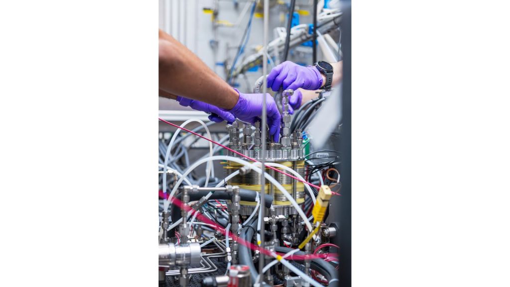 A close-up of four purple-gloved hands adjusting components inside of a system inside of a laboratory. A tangle of colorful wires and tubes extend from a gold-and-silver fuel cell in the center of the image.
