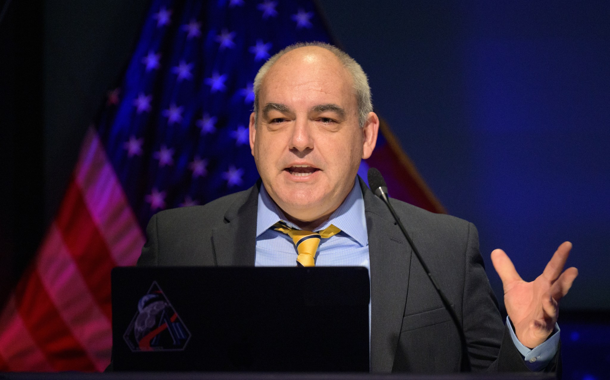 A man in a suit speaks at a podium with a microphone while gesturing with his hand. An American flag is visible in the background, and a laptop with a mission-style emblem sticker sits in front of him.