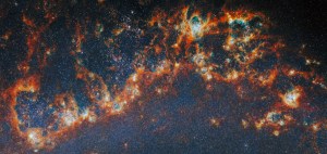A large, long portion of one of the spiral arms in galaxy M51. Red-orange, clumpy filaments of gas and dust that stretch in a chain from left to right comprise the arm. Shining cyan bubbles light up parts of the gas clouds from within, and gaps expose bright star clusters in these bubbles as glowing white dots. The whole image is dotted with small stars. A faint blue glow around the arm colors the otherwise dark background.