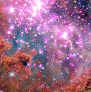 Scores of gleaming stars ringed in neon pink stretch across the frame, highlighting a cluster where stars are between one and three million years old. Brick-orange dust clouds along the bottom edge illustrate the raw materials of this active stellar nursery.