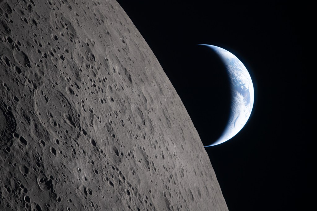 View of the Moon’s cratered surface photographed by the Artemis II crew during the mission, with a partially illuminated Earth visible beyond the lunar horizon against the blackness of space.