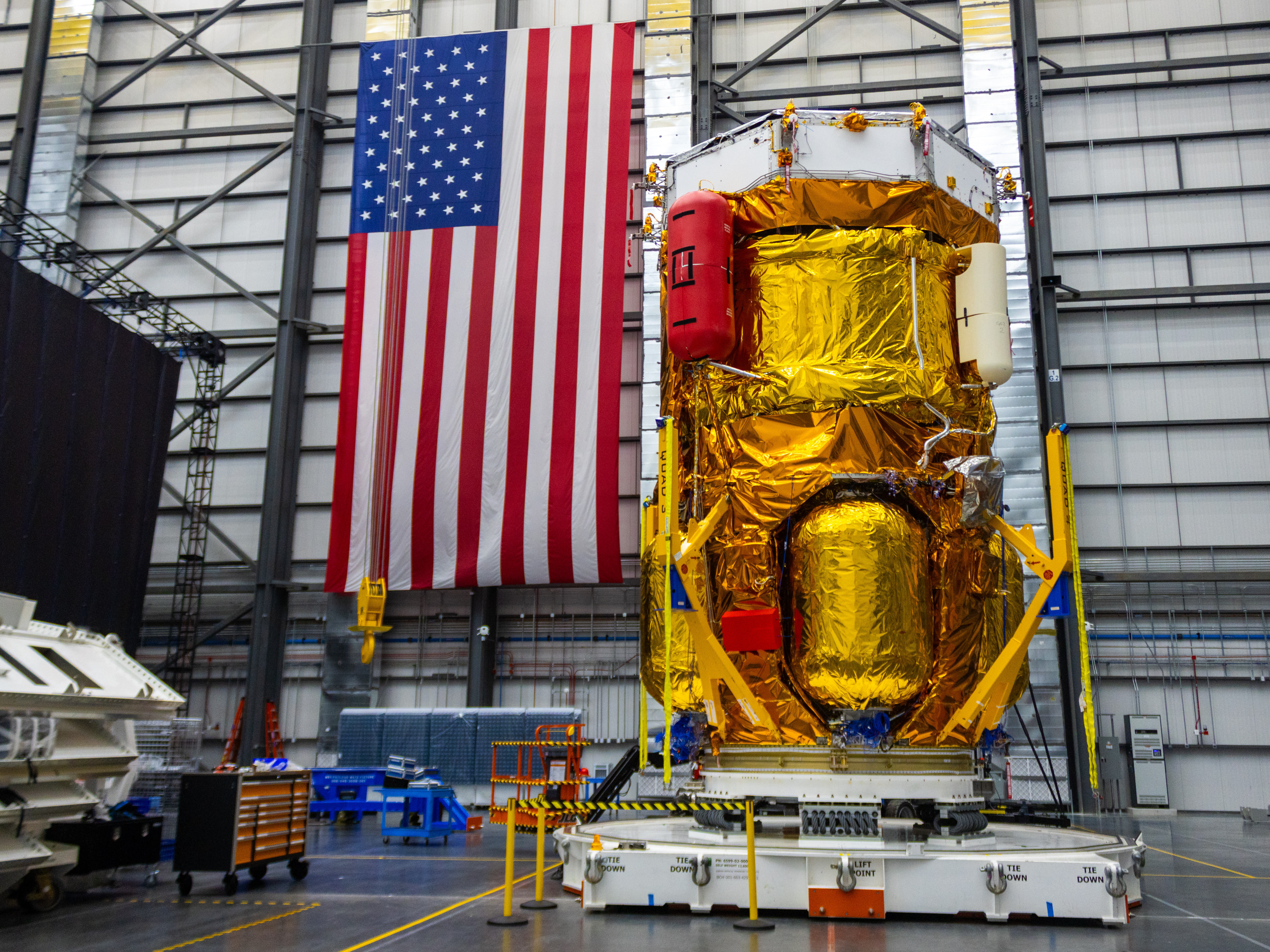 Blue Origin’s Blue Moon Mark 1 lunar lander, covered in gold thermal insulation, stands vertically inside a large processing facility beside a large American flag.