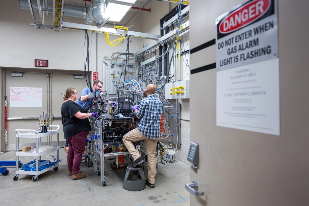 Three researchers reach inside a system filled with tubes, wires, and hardware inside a laboratory. A fuel cell sits on a white multi-tiered wheeled cart to their left along with boxes, wires, and other equipment. In the foreground is a open door with a sign that says 