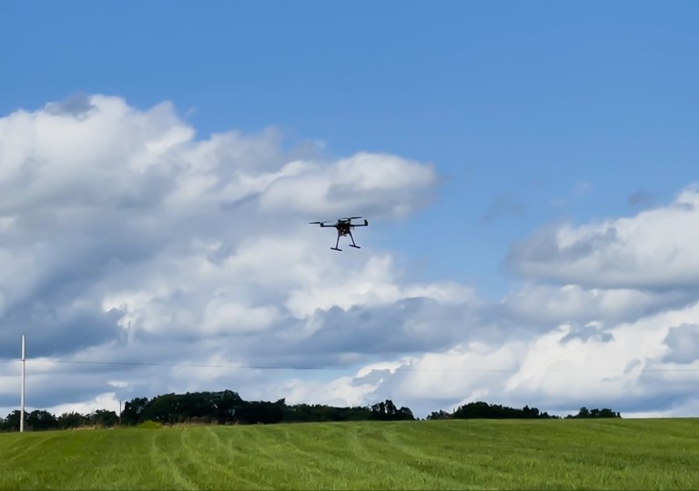 A drone with four helicopter-like blades hovers over a rural green field amidst a bright partly cloudy sky.