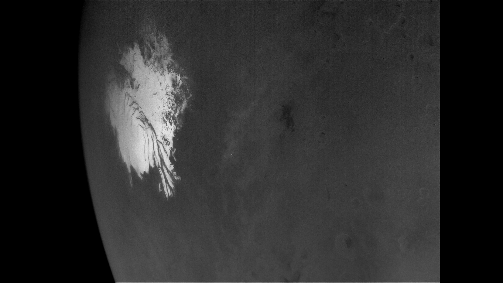 A close-up, grayscale image of the Martian surface, highlighting the intricate, swirling edges and dark troughs of a bright white polar ice cap next to surrounding plains textured with faint craters.