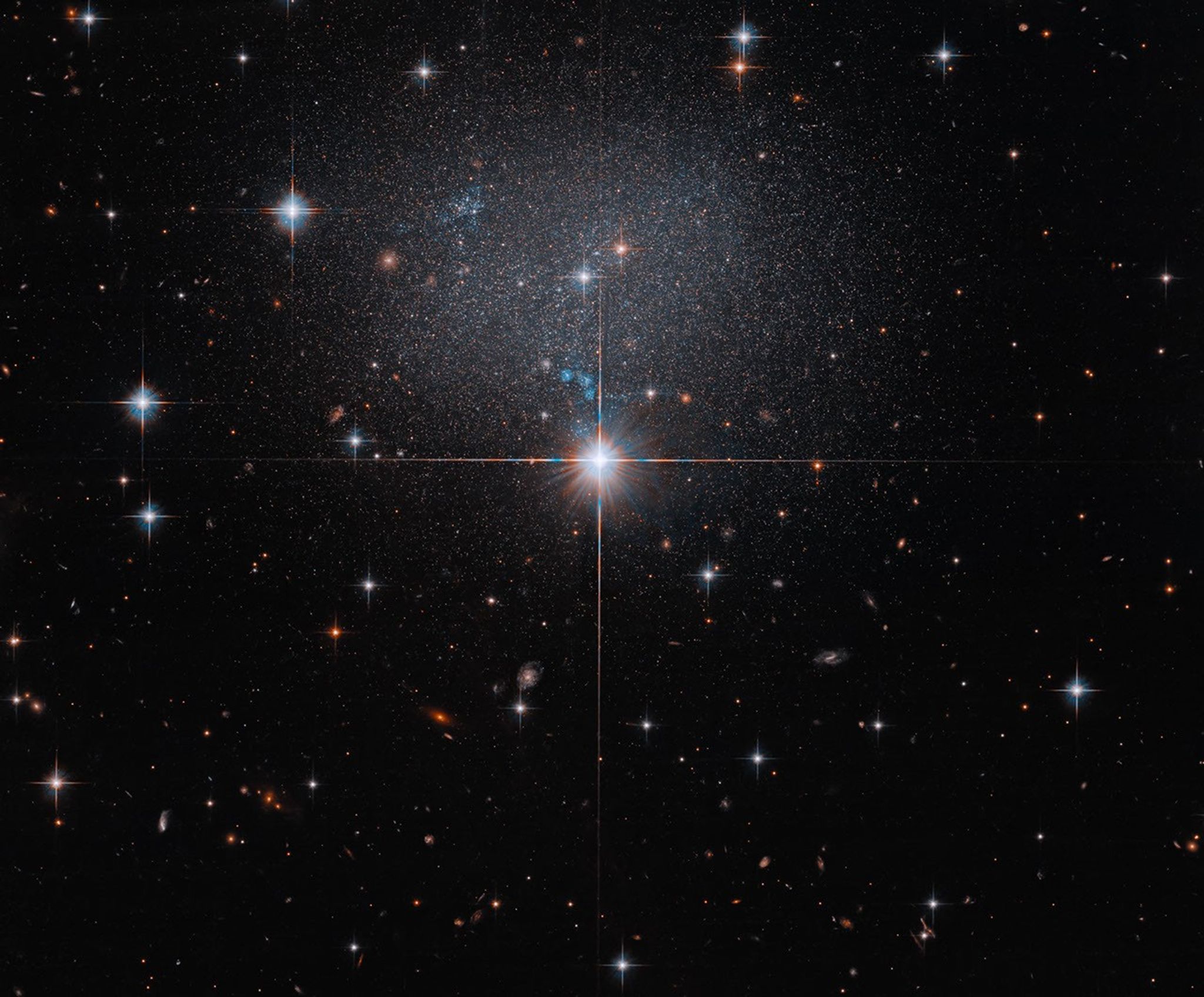 A star in the foreground shines brightly with four diffraction spikes. Above this star is a diffuse and faint dwarf irregular galaxy. The galaxy itself has bright blue and red stars.
