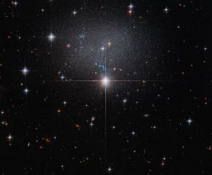 A star in the foreground shines brightly with four diffraction spikes. Above this star is a diffuse and faint dwarf irregular galaxy. The galaxy itself has bright blue and red stars.