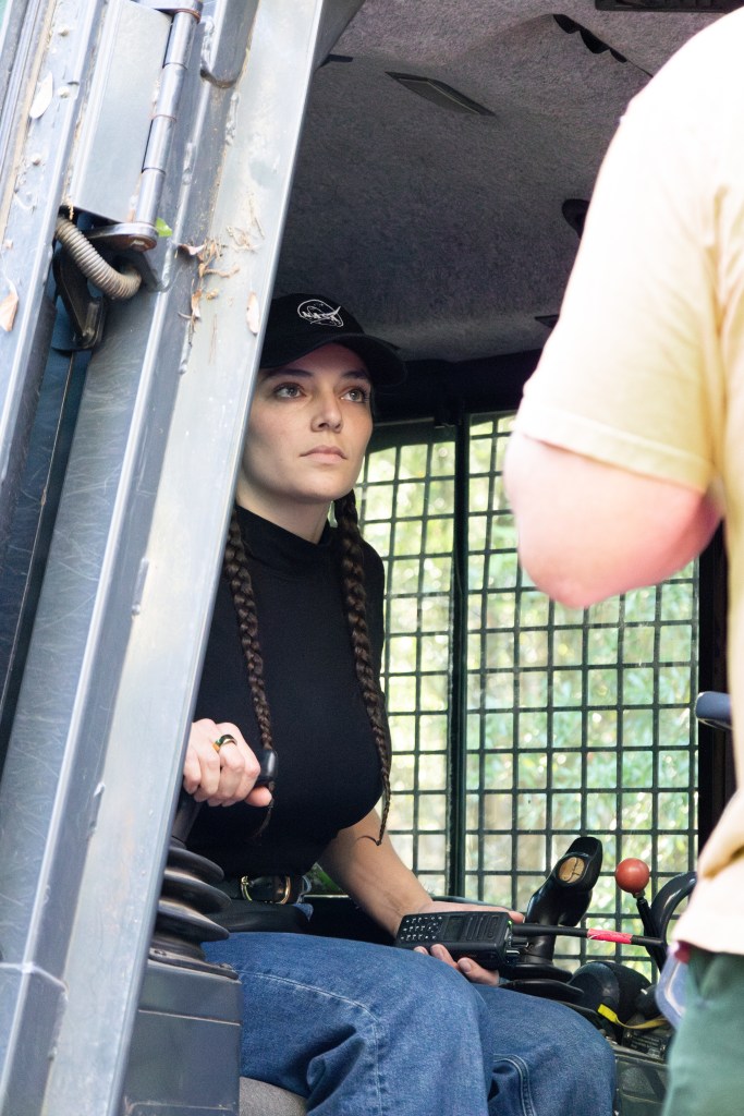 A woman wearing a black shirt, black baseball cap, and blue jeans sits inside the cab of a bulldozer looking at someone out of the screen. In her left hand is a black radio, and her right hand is on a lever.