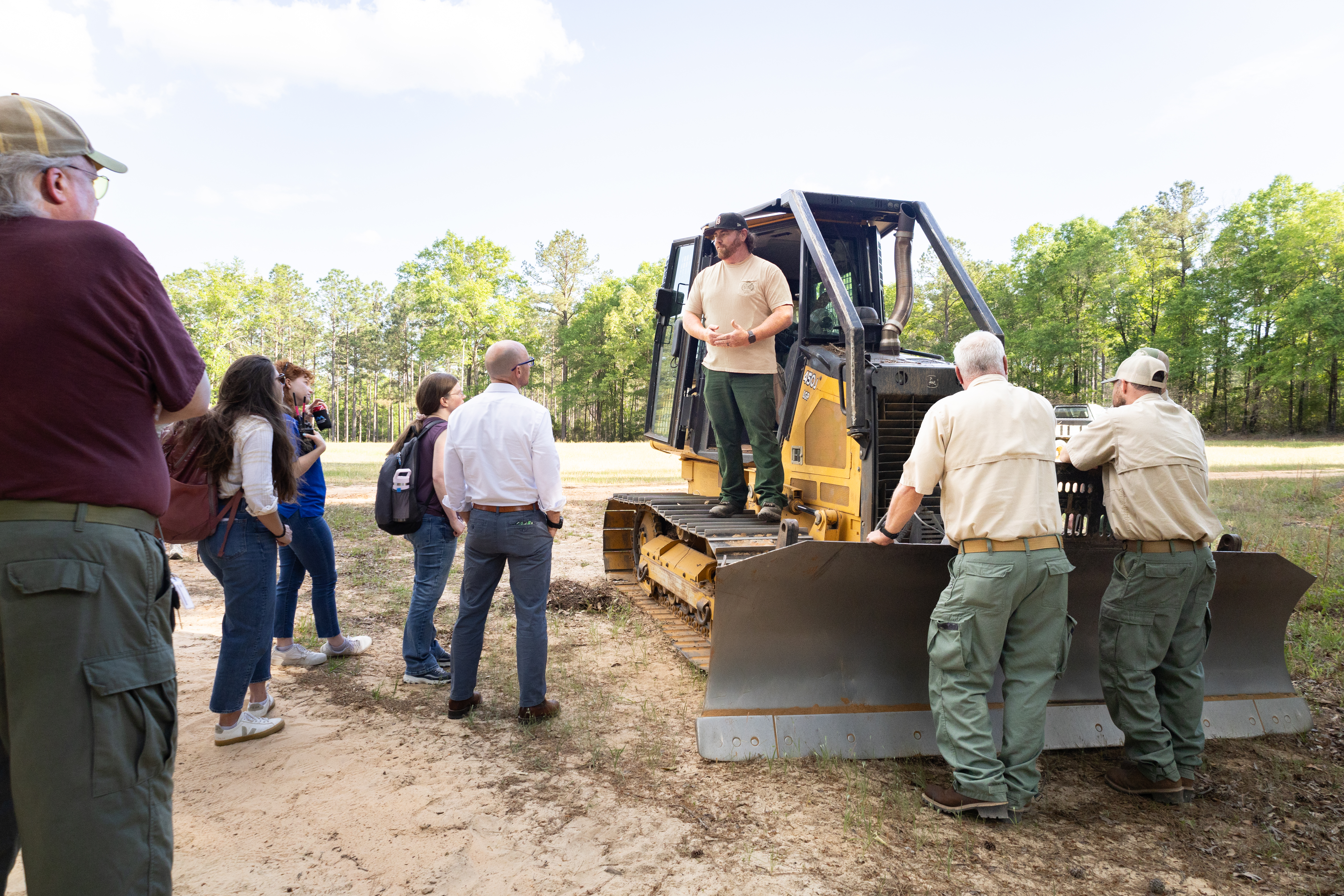 A man wearing a tan shirt, olive green pants, and a black baseball cap stands on the wheel treads of a black and yellow bulldozer. A group of men and women stand on the left of the frame, looking at the man in pink. two men in cream shirts and green pants leans on the blade of the dozer, also looking at the man in pink. In the background is a forest of trees.