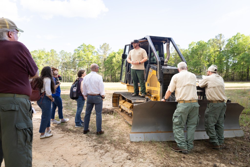 A man wearing a tan shirt, olive green pants, and a black baseball cap stands on the wheel treads of a black and yellow bulldozer. A group of men and women stand on the left of the frame, looking at the man in pink. two men in cream shirts and green pants leans on the blade of the dozer, also looking at the man in pink. In the background is a forest of trees.