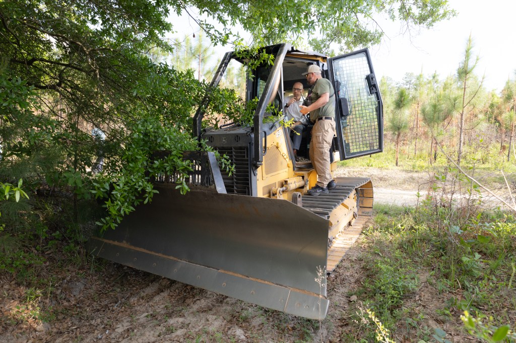 A yellow and black bulldozer sits on a dirt path next to a bushy green tree. A bald man in glasses and a white button up sits in the cab in the door open. A man in an olive green shirt and khaki pants stands on the wheel treads, leaning into the cab.