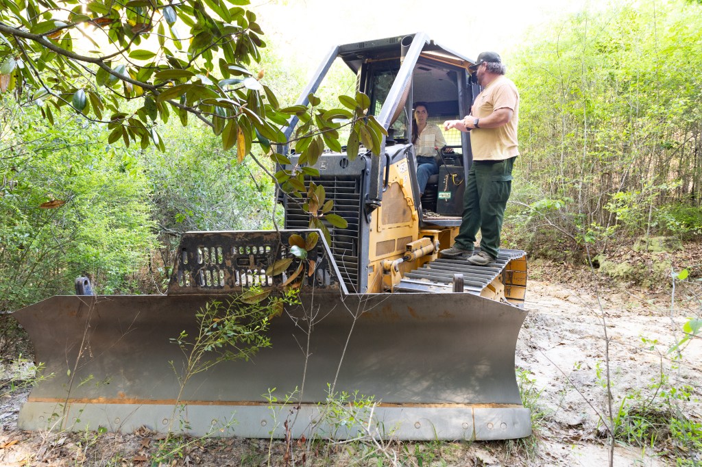 A black and yellow bulldozer sits on a dirt path, inside of a green forest, facing towards the camera. A woman in a white shirt and blue jeans sits in the cab, while a man in green pants and a tan shirt stands on the tread.