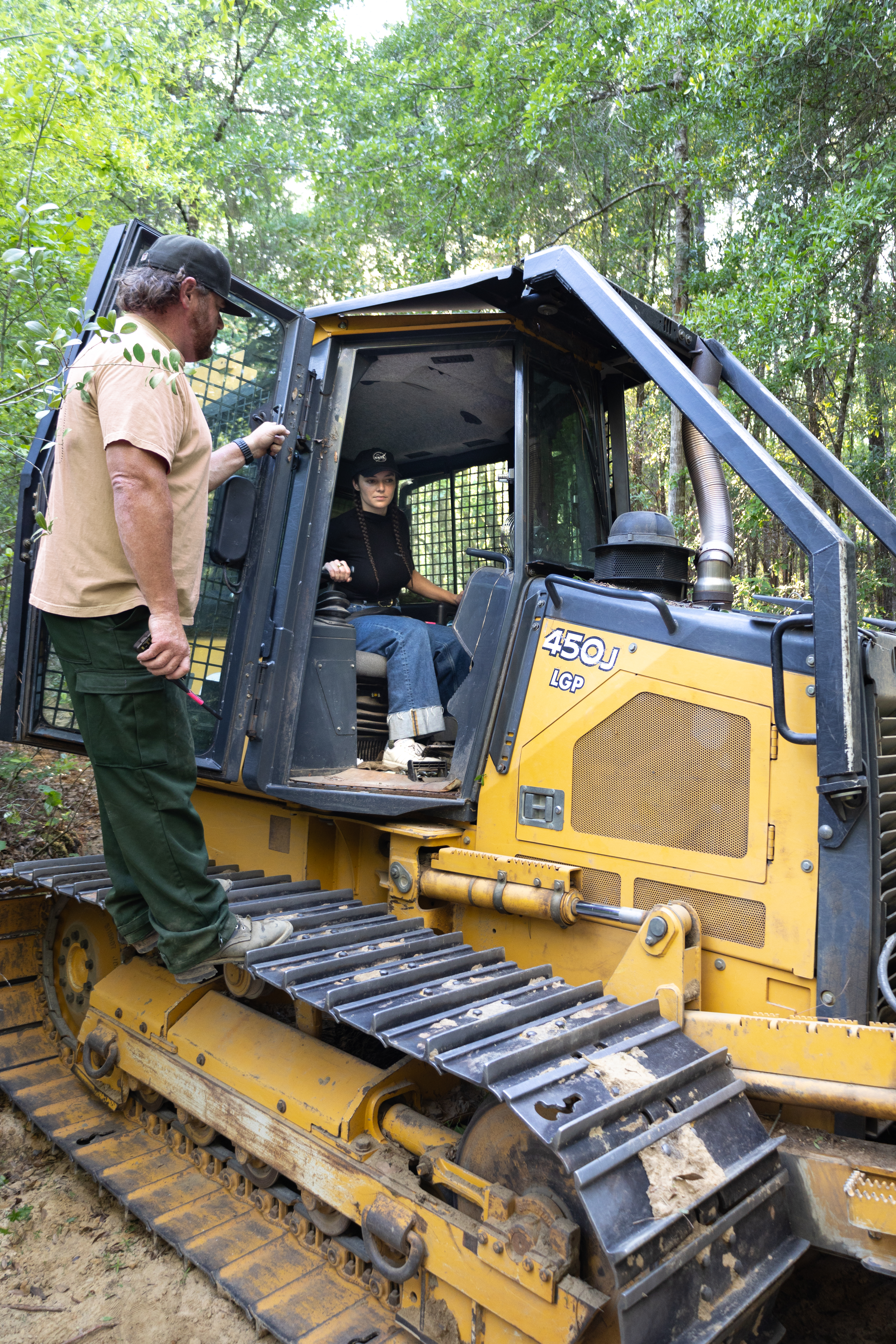 A black and yellow bulldozer sits on a dirt path, inside of a green forest, facing towards the right side of the frame. A woman in a black shirt and blue jeans sits in the cab, while a man in green pants and a tan shirt stands on the tread.