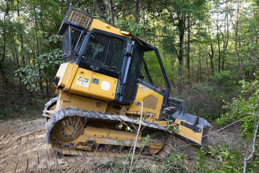 A black and yellow bulldozer sits on a dirt path, inside of a green forest. The dozer is facing from towards the right of the frame.