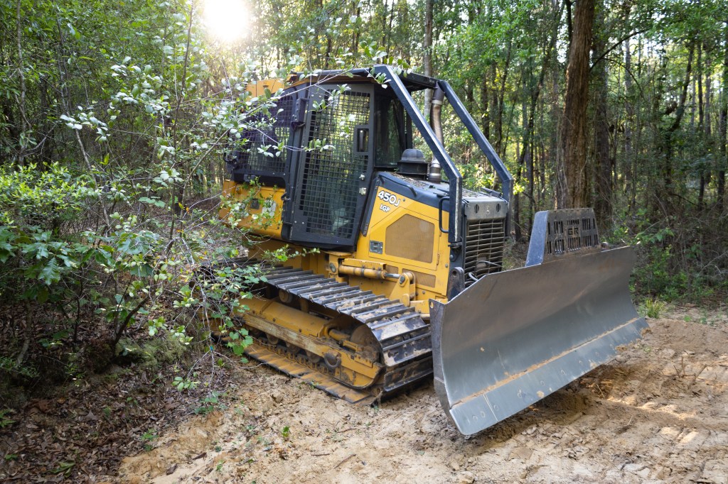 A black and yellow bulldozer sits on a dirt path, inside of a green forest. The dozer is facing from towards the right of the frame.