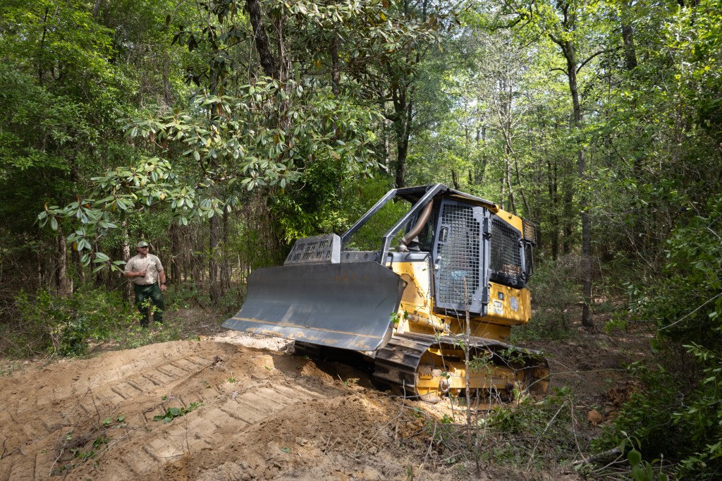 A yellow and black bulldozer climbs a small dirt hill in a forest of tall green trees, driving from the left of the frame to the right. Past the bulldozer, a man in a tan shirt and green pants stands in the trees, holding a radio.