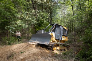 A yellow and black bulldozer climbs a small dirt hill in a forest of tall green trees, driving from the left of the frame to the right. Past the bulldozer, a man in a tan shirt and green pants stands in the trees, holding a radio.
