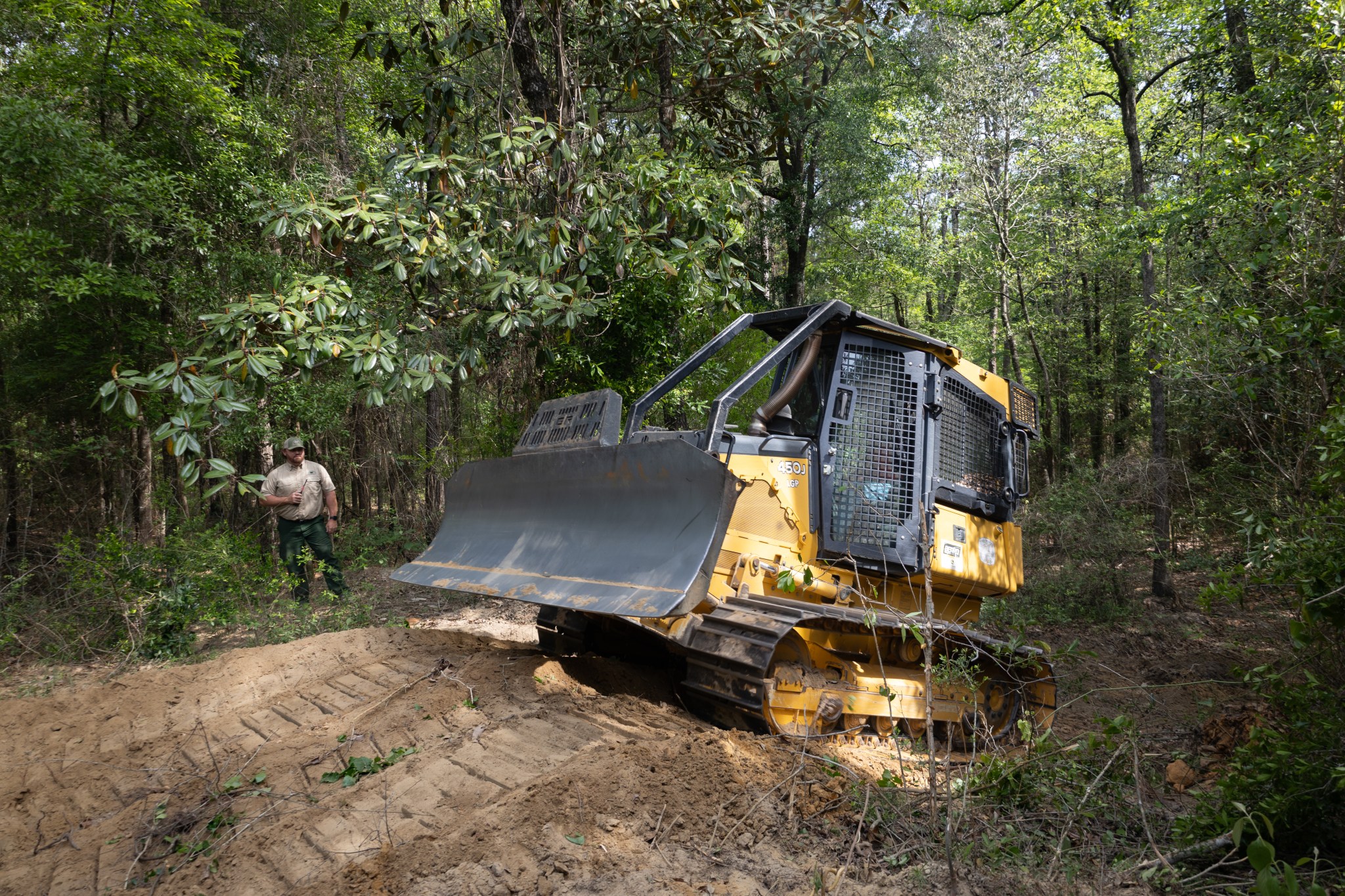 A yellow and black bulldozer climbs a small dirt hill in a forest of tall green trees, driving from the left of the frame to the right. Past the bulldozer, a man in a tan shirt and green pants stands in the trees, holding a radio.