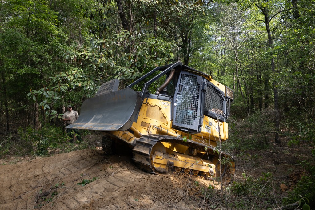 A yellow and black bulldozer climbs a small dirt hill in a forest of tall green trees, driving from the left of the frame to the right. Past the bulldozer, a man in a tan shirt and green pants stands in the trees, holding a radio.