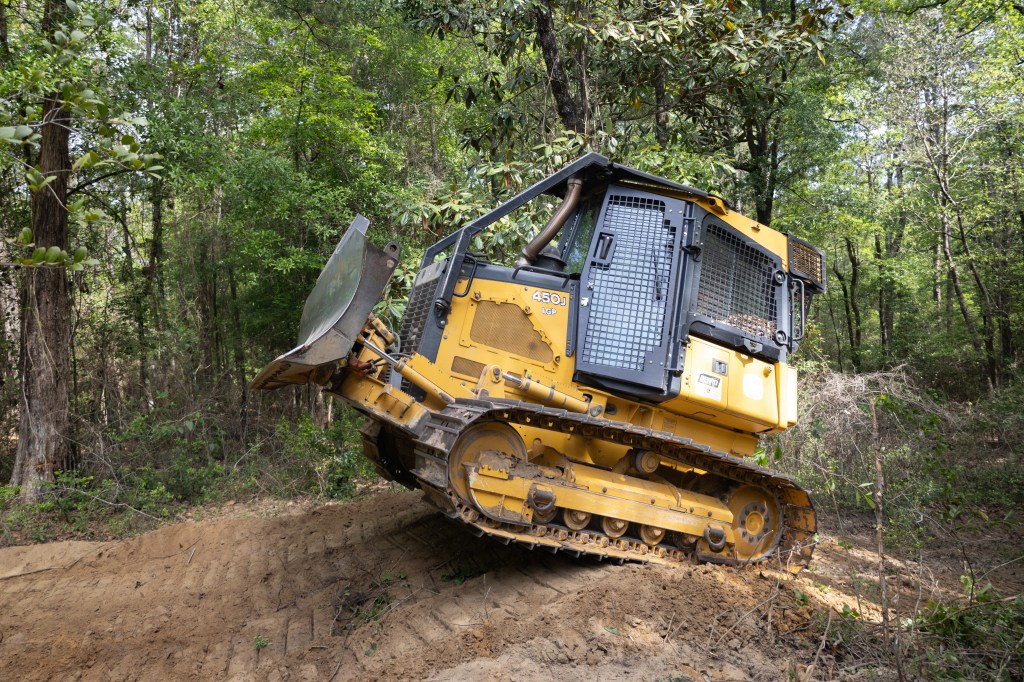 A yellow and black bulldozer climbs a small dirt hill in a forest of tall green trees, driving from the left of the frame to the right.