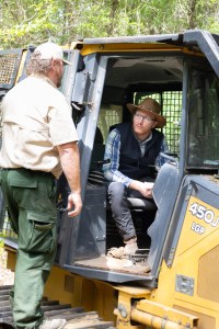 A man wearing a blue shirt, black vest, straw hat, and black jeans sits inside the cab of a bulldozer. A man in green pants, tan shirt, and tan baseball cap, holding a radio, stands just outside the cab leaning in.