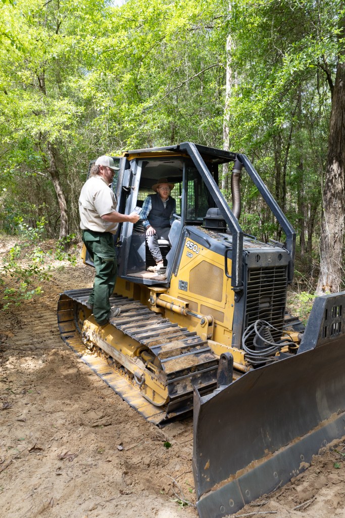 A black and yellow bulldozer sits on a dirt path, inside of a green forest, facing towards the right side of the frame. A man in a blue shirt, black vest, and black jeans sits in the cab, while a man in green pants and a tan shirt stands on the tread.