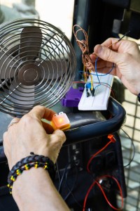 Hands belonging to someone offscreen hold sensor components onto a small black bar. In her left hand is a small yellow box about an inch big, with a bright LED light turned on in the middle. In her right hand is a jumble of wires. In front of the sensor components is a small metal fan.