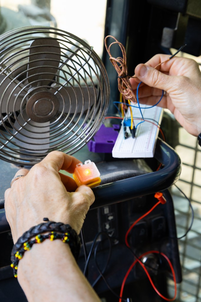 Hands belonging to someone offscreen hold sensor components onto a small black bar. In her left hand is a small yellow box about an inch big, with a bright LED light turned on in the middle. In her right hand is a jumble of wires. In front of the sensor components is a small metal fan.