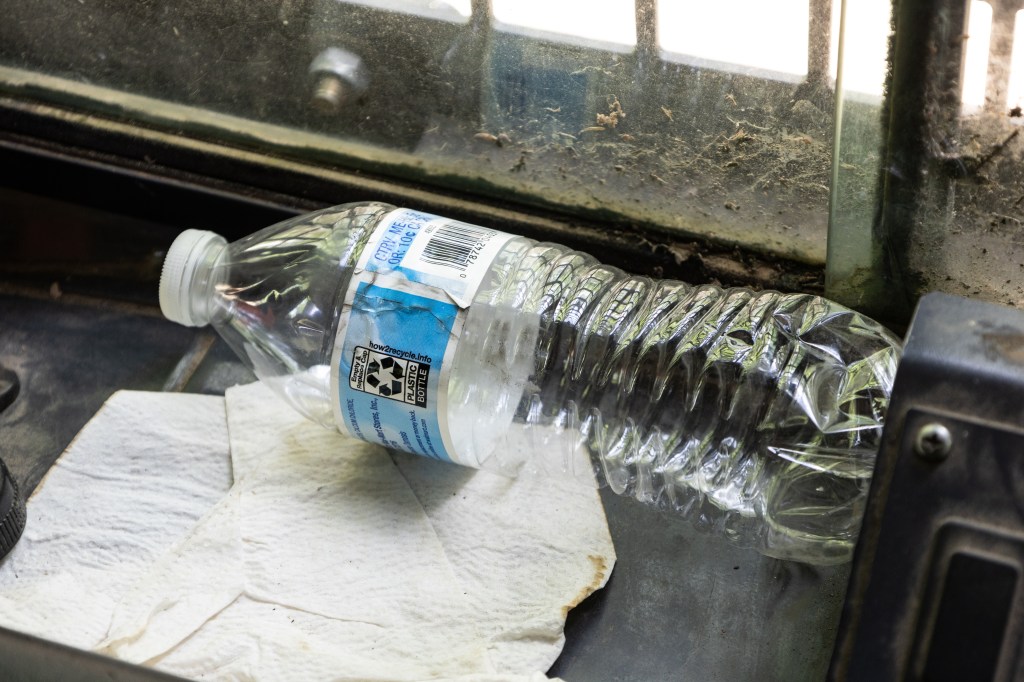 An empty plastic water bottle with a light blue label sits on a piece of metal and a discarded cream colored napkin, up against a dirty window.