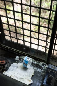 An empty plastic water bottle with a light blue label sits on a piece of metal and a discarded cream colored napkin, up against a dirty window covered in a cross-lattice of bars.