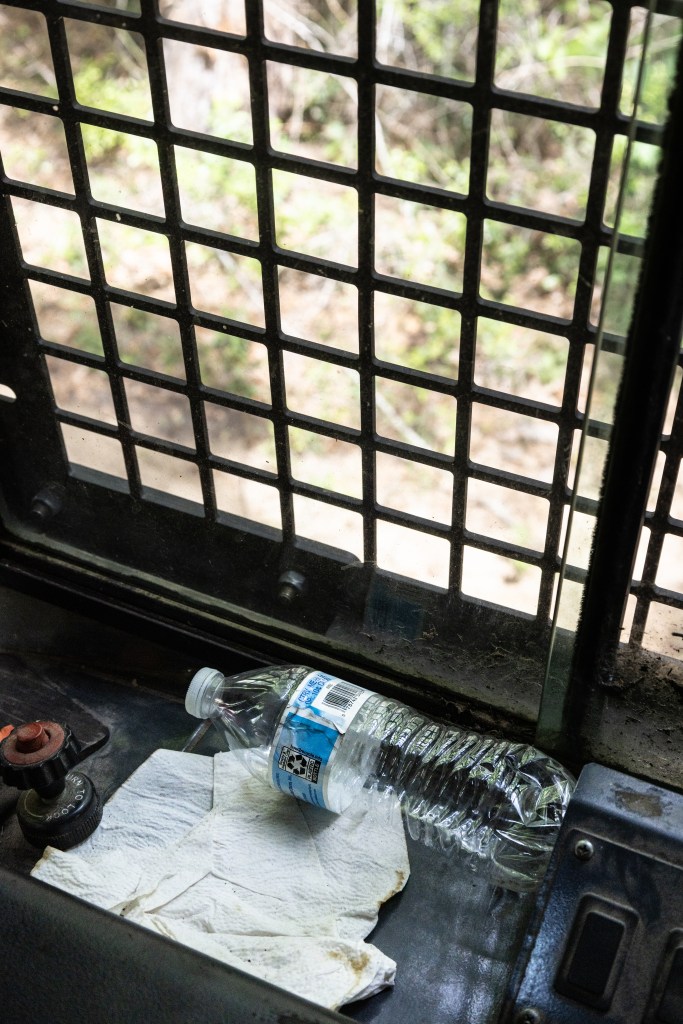 An empty plastic water bottle with a light blue label sits on a piece of metal and a discarded cream colored napkin, up against a dirty window covered in a cross-lattice of bars.