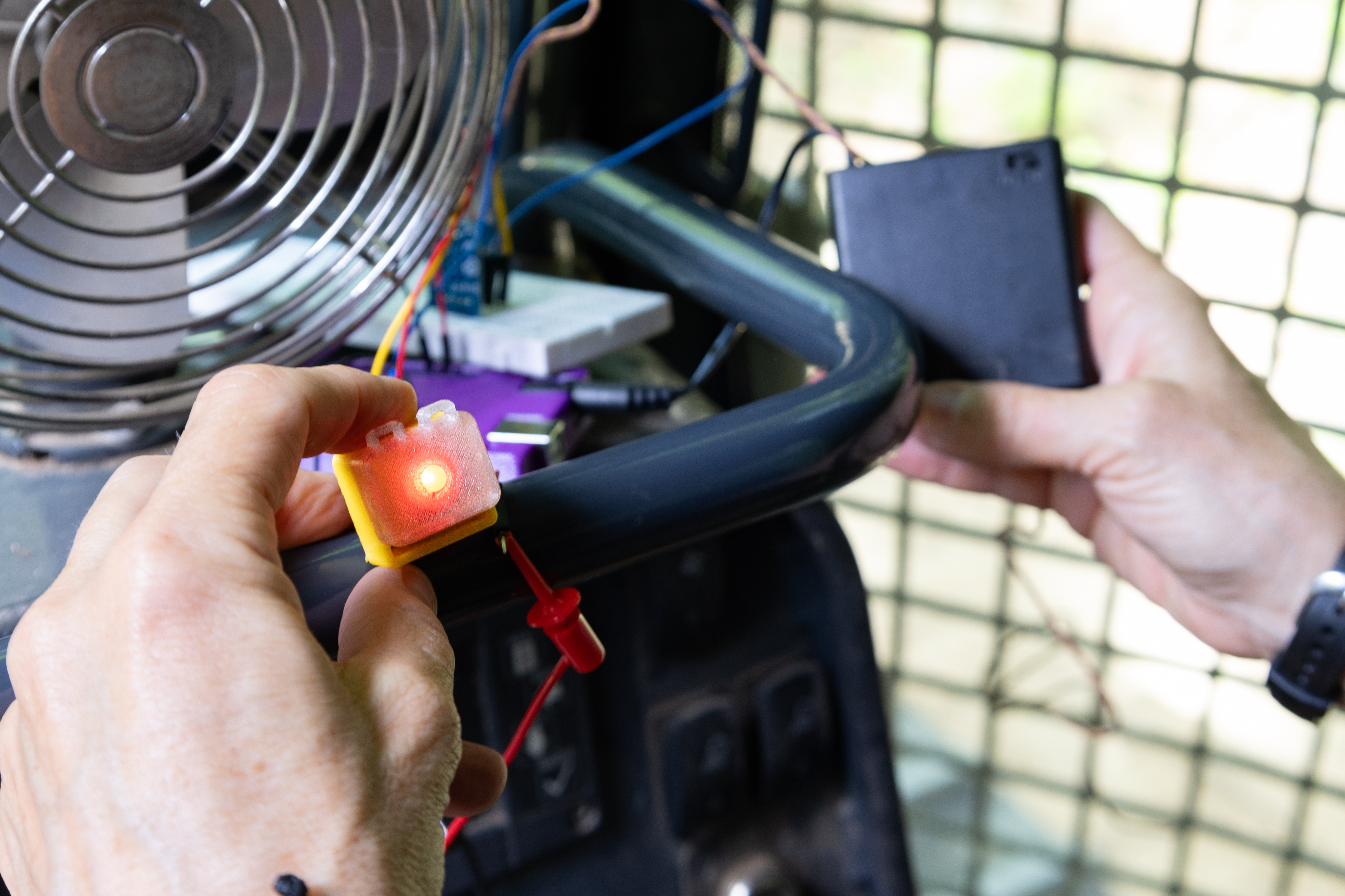 Hands belonging to someone offscreen hold sensor components onto a small black bar. In her left hand is a small yellow box about an inch big, with a bright LED light turned on in the middle. In her right hand is a black box with a small switch. In front of the sensor components is a small metal fan.