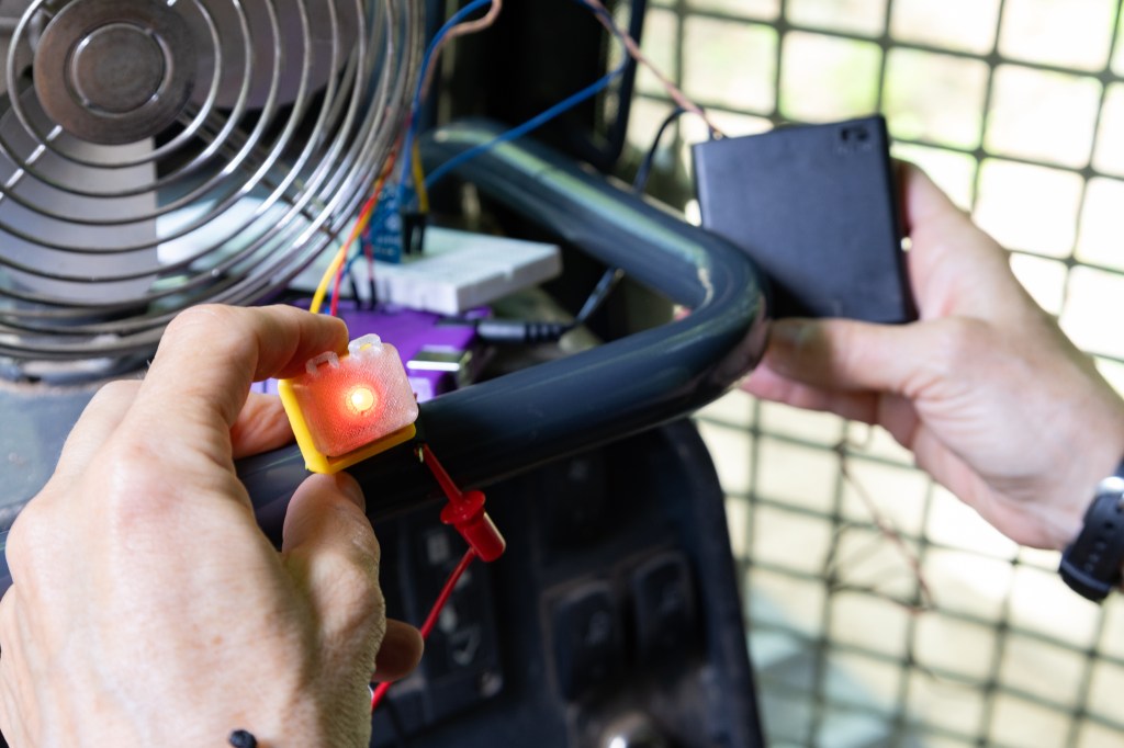 Hands belonging to someone offscreen hold sensor components onto a small black bar. In her left hand is a small yellow box about an inch big, with a bright LED light turned on in the middle. In her right hand is a black box with a small switch. In front of the sensor components is a small metal fan.