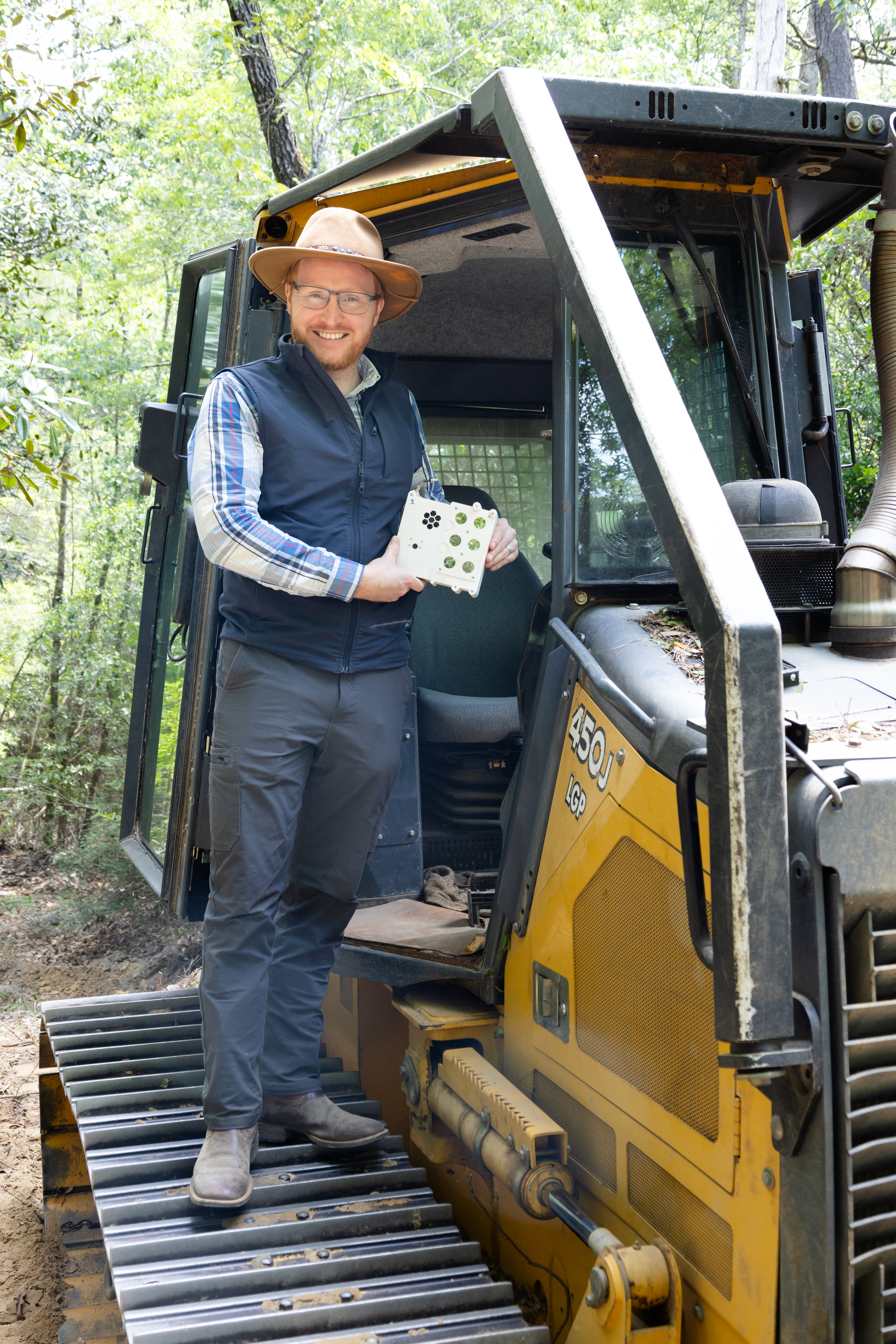 A man in dark gray jeans, a blue checkered long-sleeve, black vest, and straw hat stands on the tread of a black and yellow bulldozer, holding a white sensor box the size of a lunchbox and smiling at the camera.
