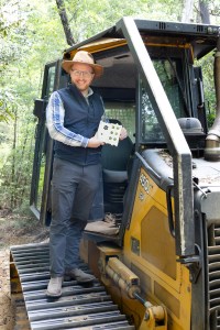 A man in dark gray jeans, a blue checkered long-sleeve, black vest, and straw hat stands on the tread of a black and yellow bulldozer, holding a white sensor box the size of a lunchbox and smiling at the camera.