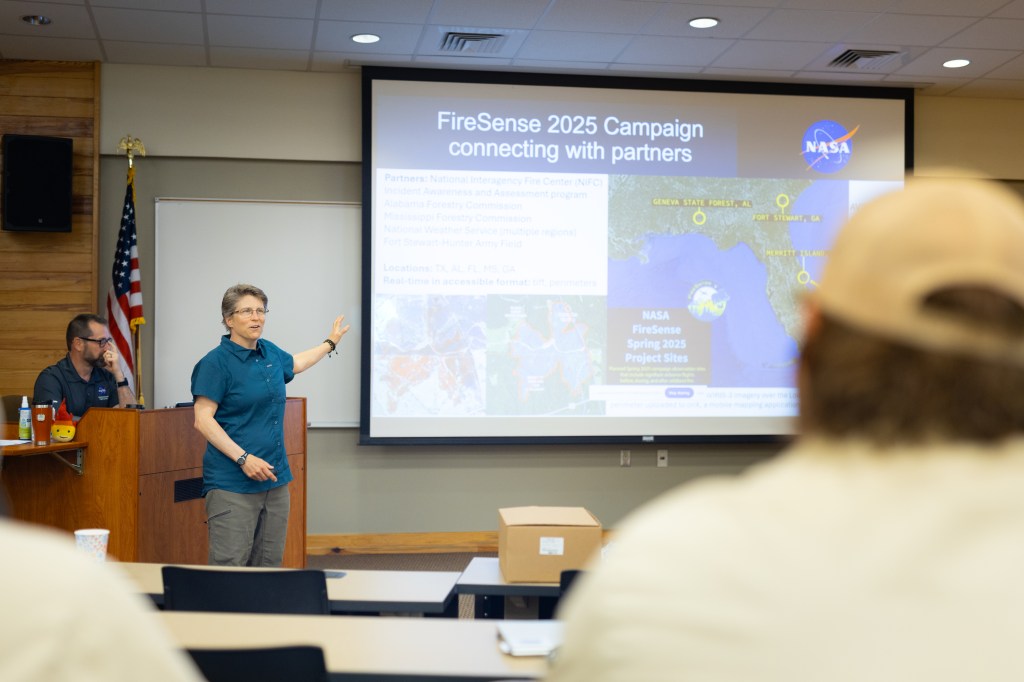 A woman in a teal t-shirt and olive green pants stands in the left of the frame, in front of an auditorium. She is gesturing at the screen behind her, which has a slide up with maps and some text. In the foreground, a few people sit with their back to the camera.