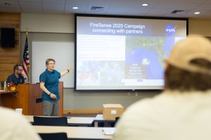 A woman in a teal t-shirt and olive green pants stands in the left of the frame, in front of an auditorium. She is gesturing at the screen behind her, which has a slide up with maps and some text. In the foreground, a few people sit with their back to the camera.