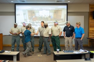 A woman in a teal t-shirt and olive green pants passes out patches to a group of men standing in a line at the front of the auditorium, most of whom are wearing olive green pants and tan t-shirts. The two men at the end are wearing a black t-shirt and tan pants, and a blue t-shirt and gray pants, respectively.