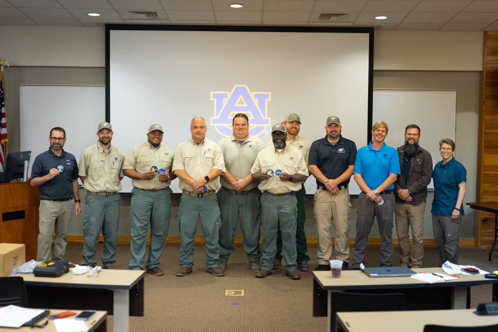 A group of 11 adults stands at the front of an auditorium, posing for a group photo. In the background is a screen displaying the Auburn University logo.