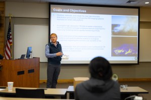 A man in dark grey pants, a blue long sleeve, and a black vest stands in the left of the frame, in front of an auditorium. She is gesturing at the screen behind her, which has a slide up with maps and some text. In the foreground, a few people sit with their back to the camera.