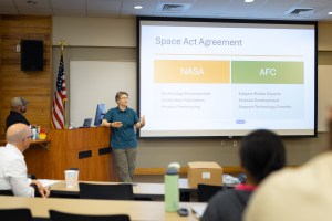A woman in a teal t-shirt and olive green pants stands in the left of the frame, in front of an auditorium. She is gesturing at the screen behind her, which has a slide up with a gold rectangle and a green rectangle. In the foreground, a few people sit with their back to the camera.