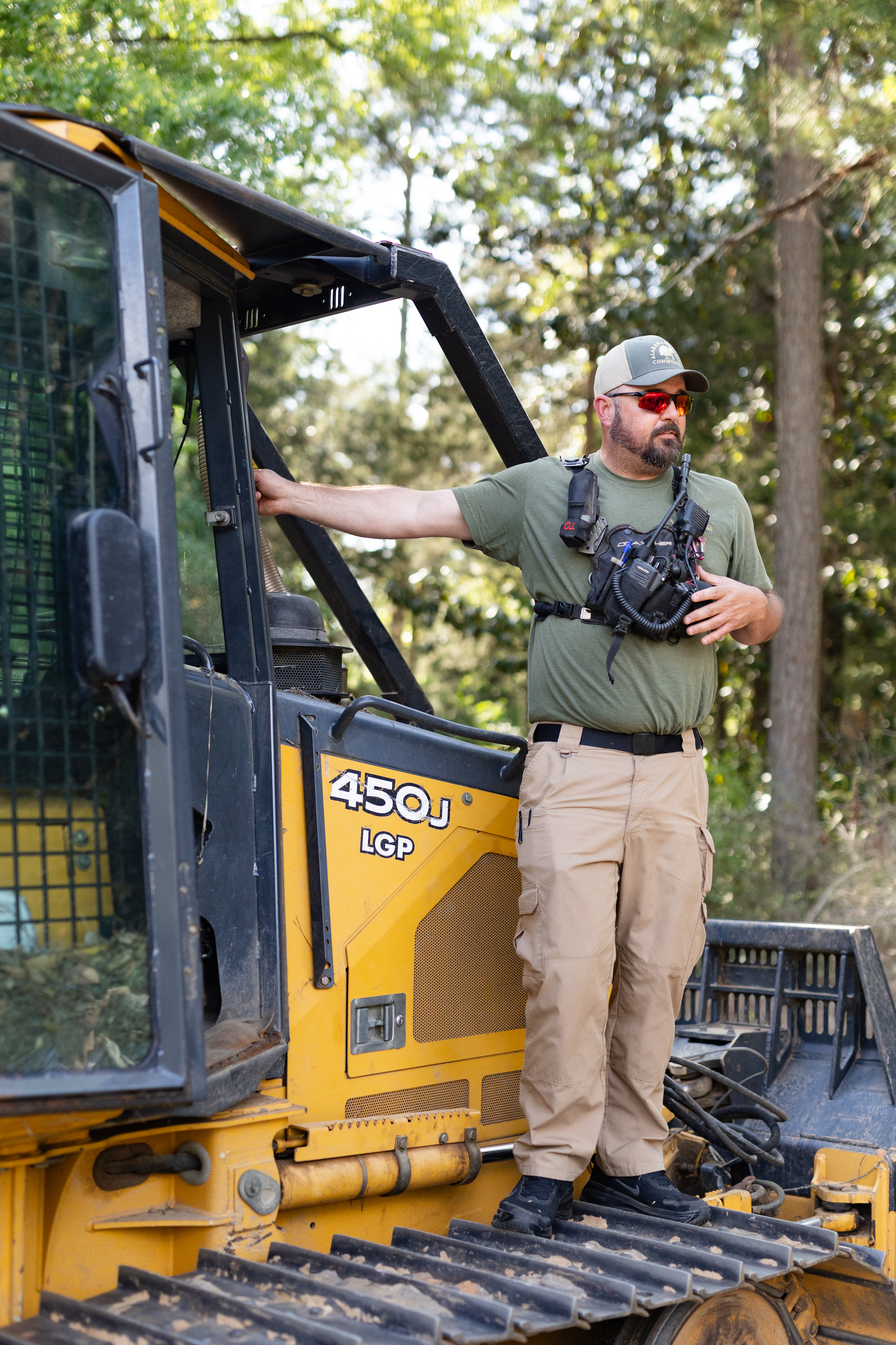 A man in an olive green shirt, tan pants, a baseball cap, red sunglasses, and a black equipment vest stands on the tread of a black and yellow bulldozer, looking off to the right of the frame.