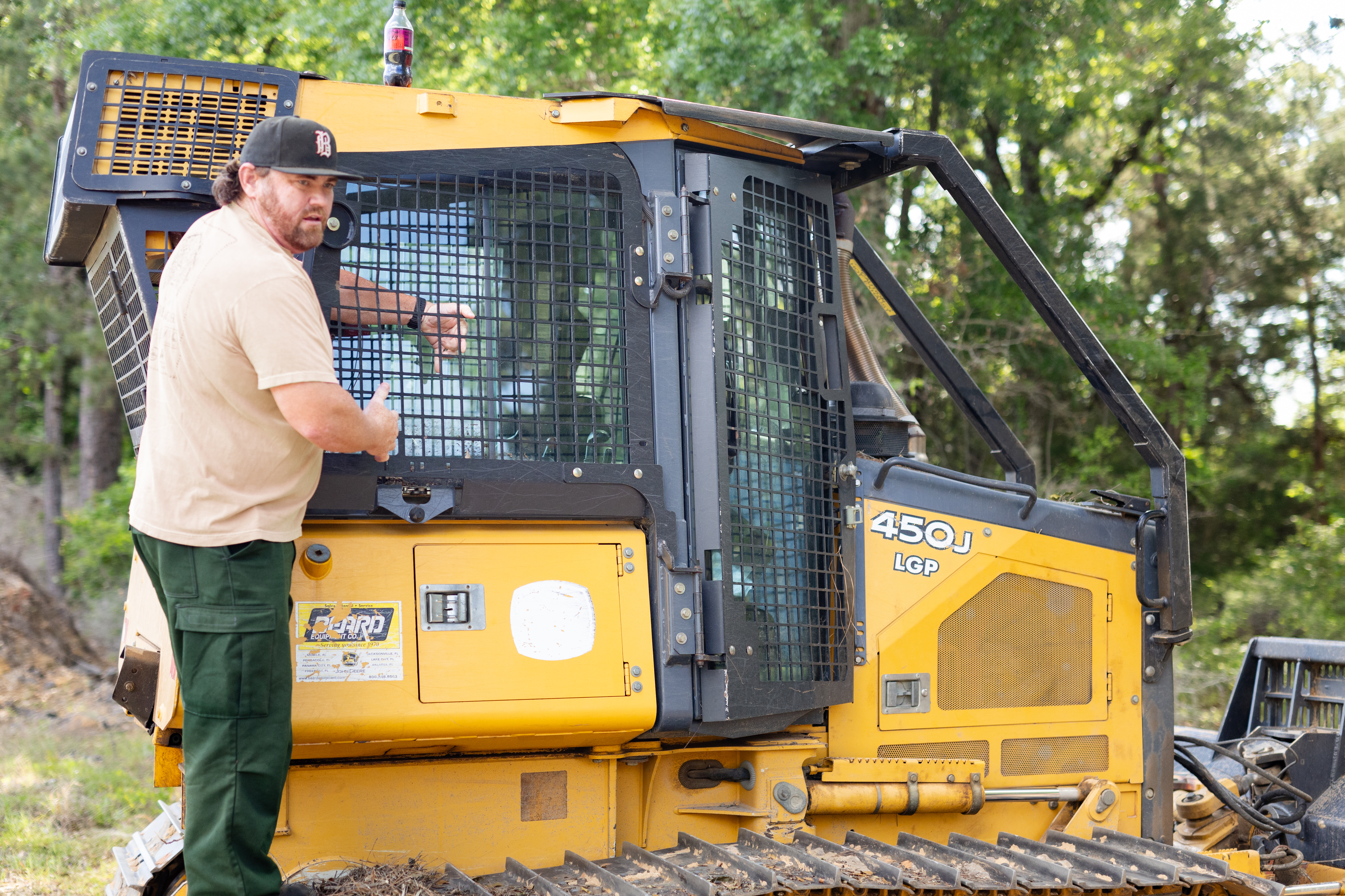 A man in olive pants, a tan shirt, and a black baseball cap stands on the tread of a black and yellow bulldozer, with his arm inside the window grate.
