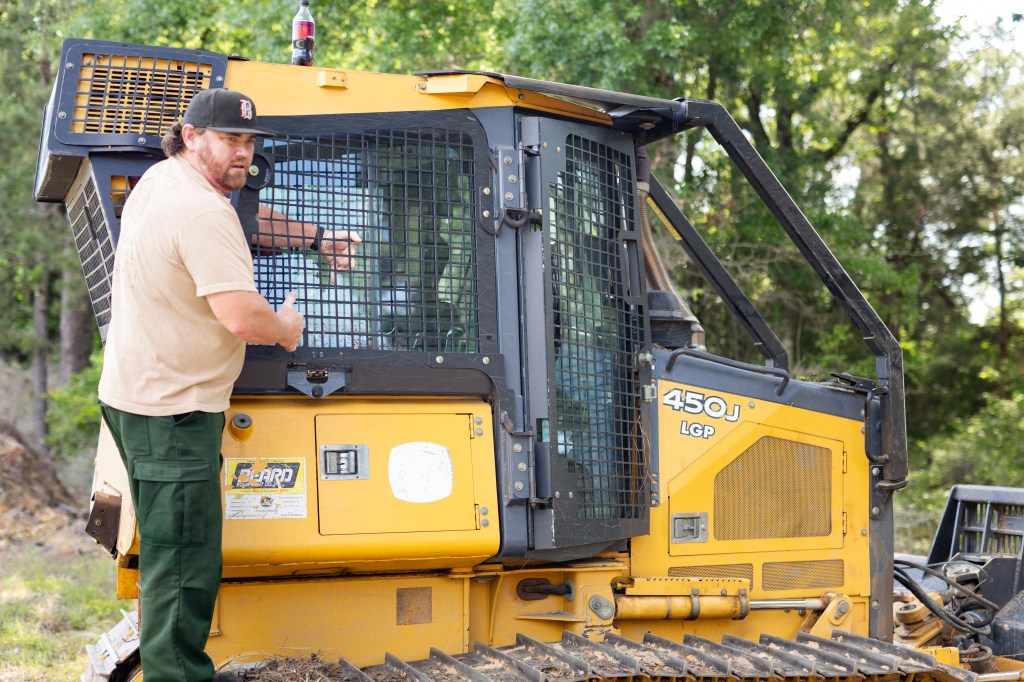 A man in olive pants, a tan shirt, and a black baseball cap stands on the tread of a black and yellow bulldozer, with his arm inside the window grate.