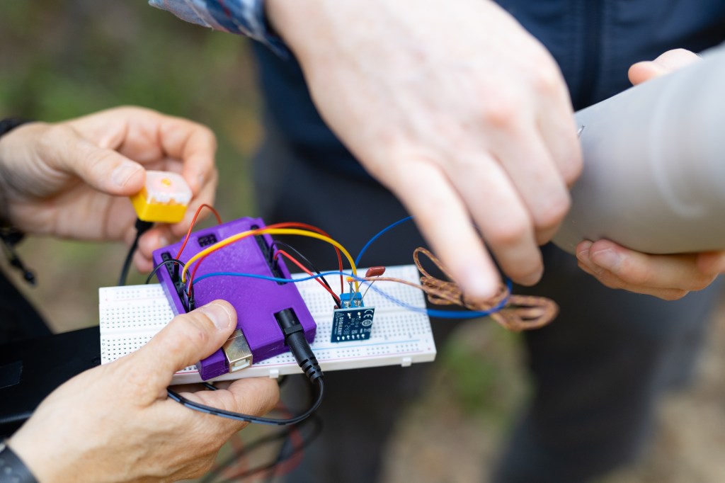 Two people's hands hold sensor components into the frame: the person on the left holds a small bright purple box, a white breadboard, a small yellow box with an LED turned on in the middle, while the person on the right holds a few of the wires against a reusable water bottle.